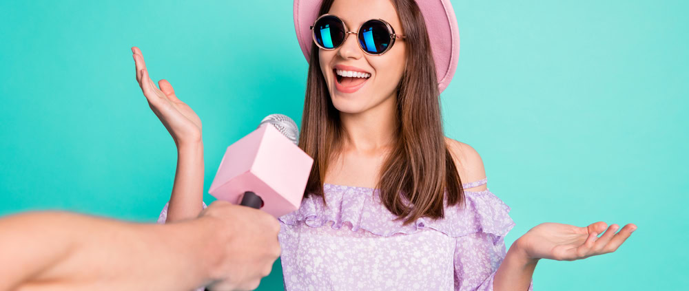 Photo of a happy young woman wearing a hat and sunglasses speaking into a microphone