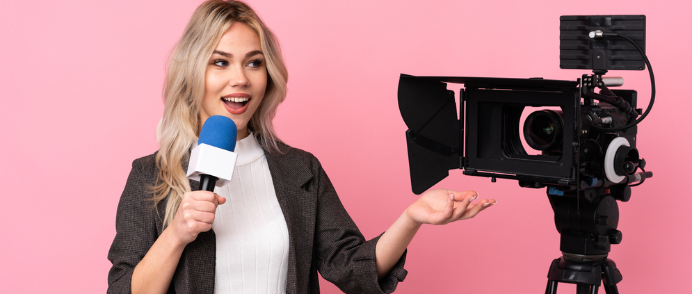 A young female reporter holding a microphone with a camera next to her.