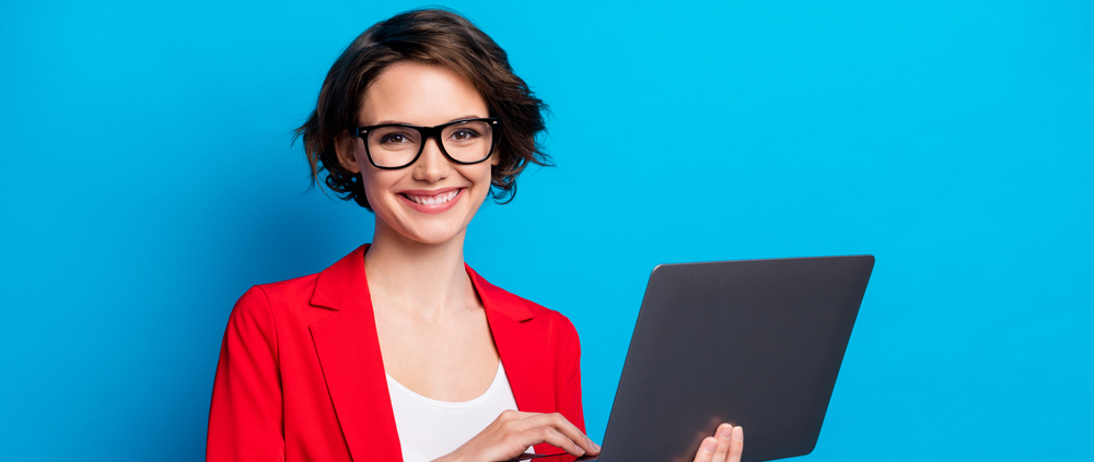 A businesswoman in a red blazer and gasses, holds a laptop and smiles at the camera.
