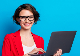 A businesswoman in a red blazer and gasses, holds a laptop and smiles at the camera.
