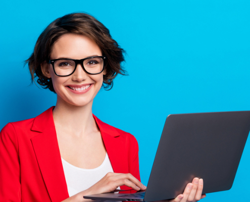 A businesswoman in a red blazer and gasses, holds a laptop and smiles at the camera.