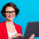 A businesswoman in a red blazer and gasses, holds a laptop and smiles at the camera.
